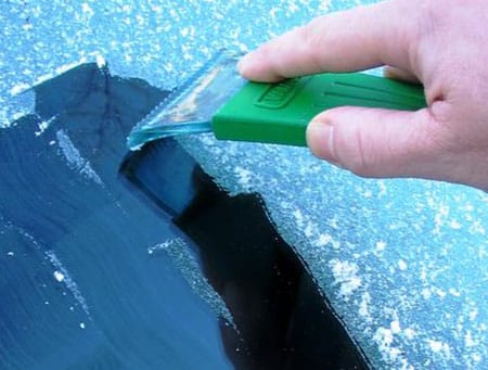 A hand using a green scraper to remove ice from a frosted car windshield.