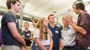 A group of people, mostly teens, listen attentively to a young man speaking about event planning in a bright indoor setting.