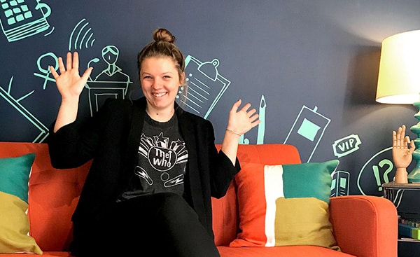 Woman smiling and waving while sitting on an orange couch, with a blue graphic wall behind her.