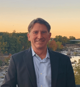 A man in a suit, possibly a 10kSB participant, stands outdoors by a river at sunset, framed by trees and a clear sky in the background.