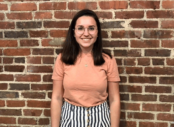 Woman in glasses and a peach t-shirt stands smiling in front of a brick wall, embodying the confidence and enthusiasm of a communications intern.