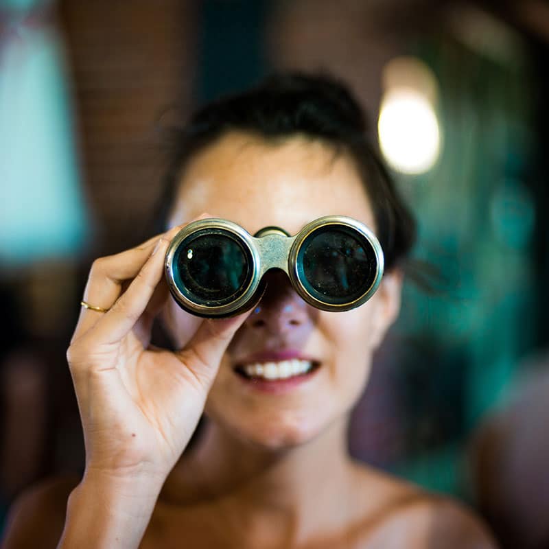 Smiling woman holding vintage binoculars up to her eyes, looking through them indoors, symbolizing the search for brand awareness.