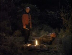 A man stands by a campfire at night, ready to communicate effectively with another person who lies on the ground nearby in a forested area.