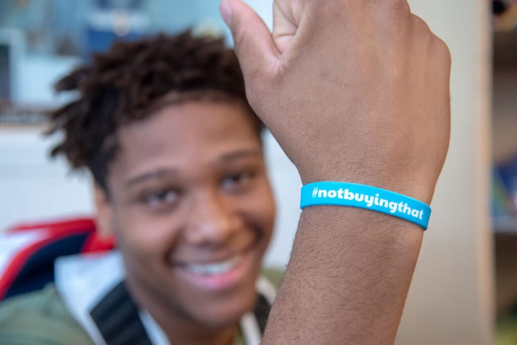 Smiling person shows a blue wristband that says #notbuyingthat in white text, supporting a youth empowerment program.