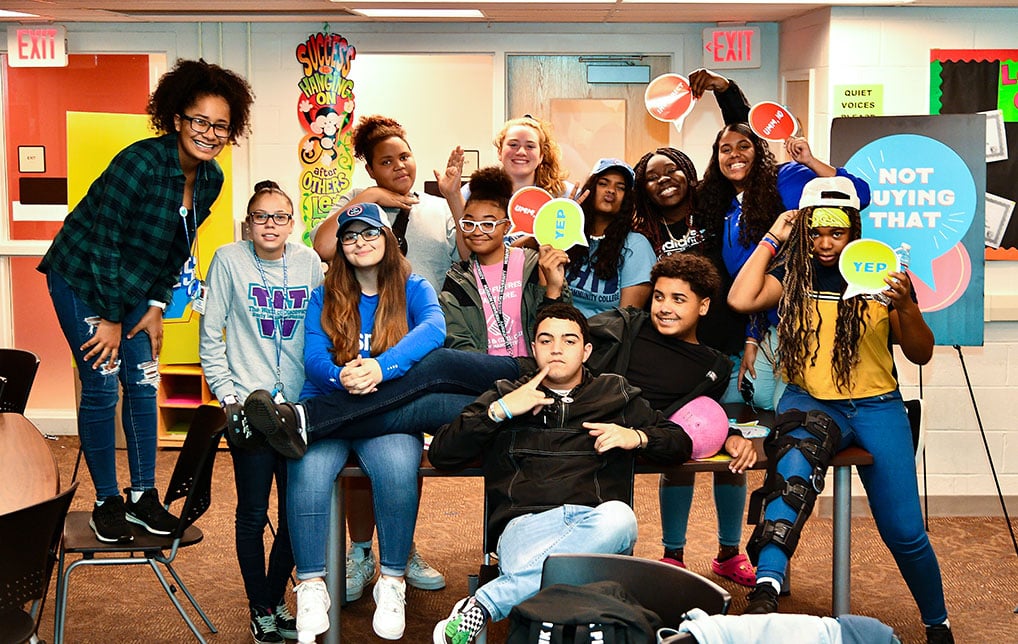 A diverse group of teens pose together indoors, smiling and holding colorful signs with positive messages as part of a youth empowerment program.
