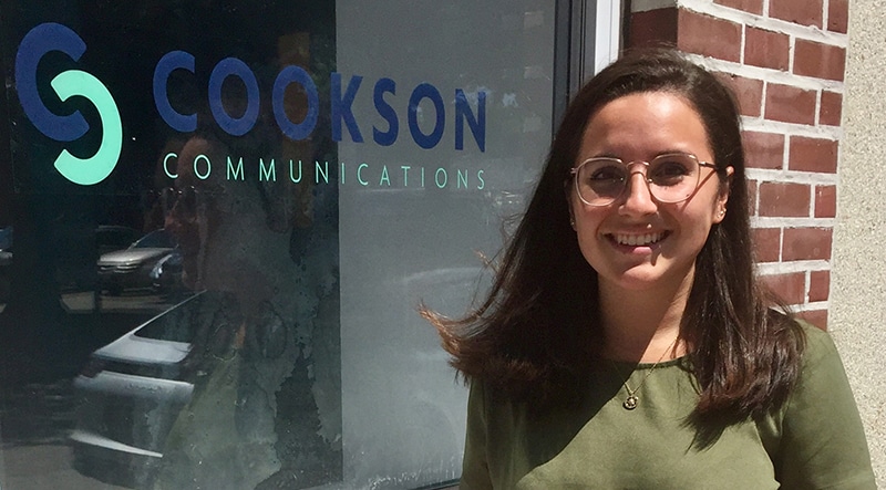 Woman with glasses smiling outside Cookson Communications office, standing next to the company logo window as a communications intern.