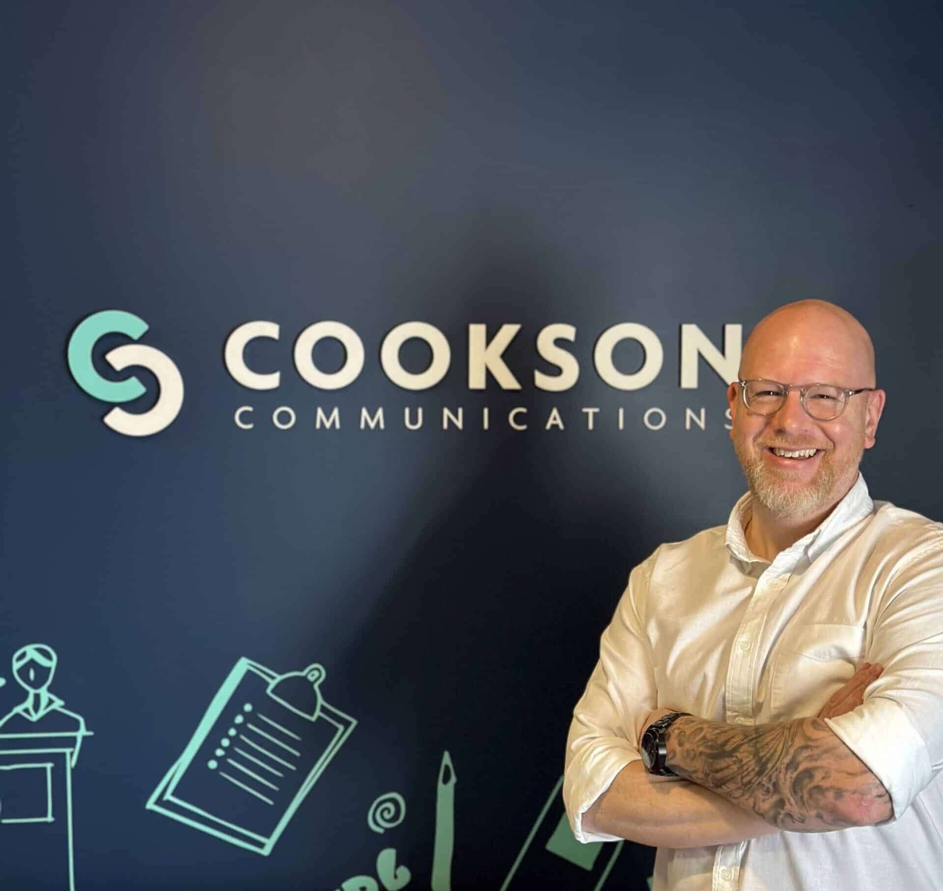Smiling man with glasses stands in front of a “Cookson Communications” sign on a blue background.