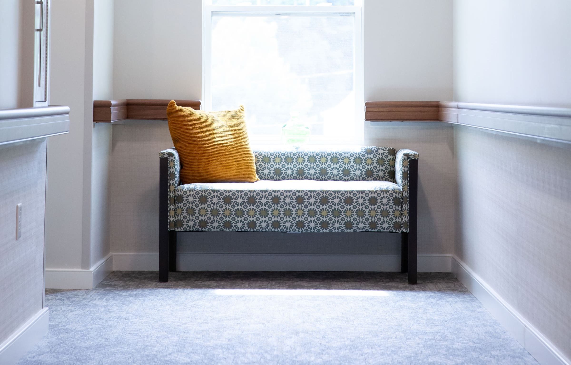 A patterned bench with a yellow pillow, part of the recent rebranding, sits by a bright window in a quiet hallway.