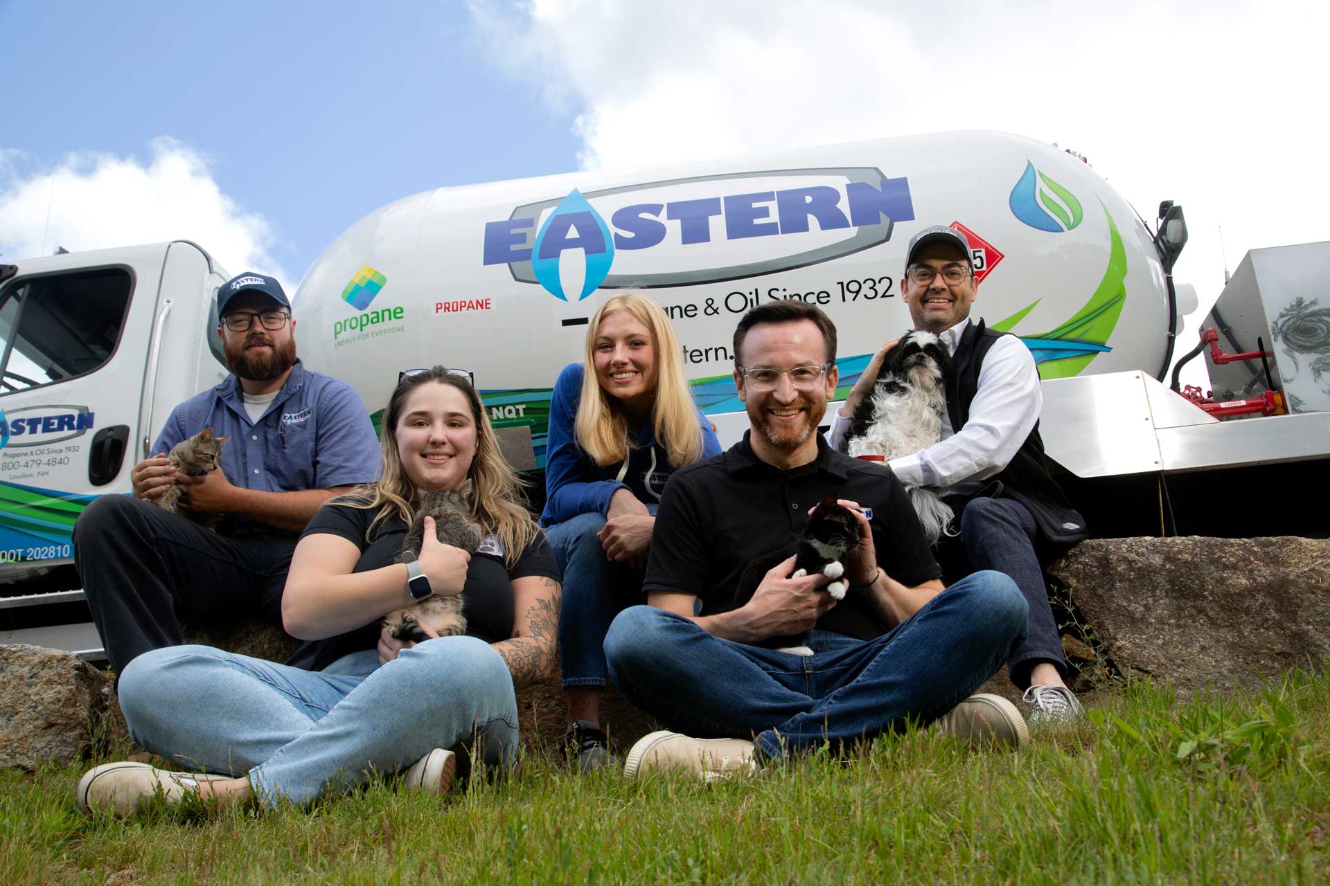 Five people sit on grass with pets in front of an Eastern propane truck, smiling at the camera.