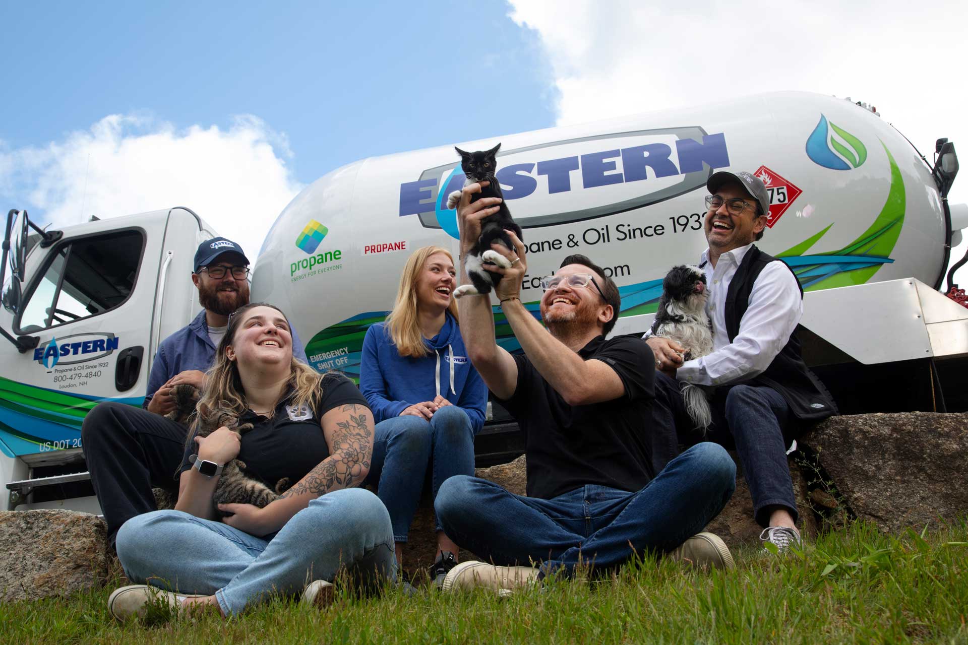 Five smiling people sit on grass in front of a propane truck, holding and playing with two cats and a dog.