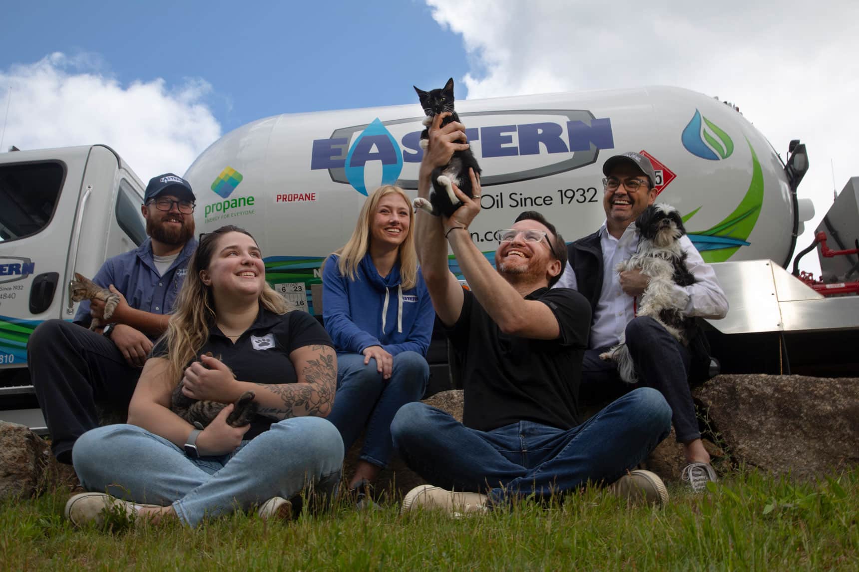 Five smiling people sit on grass with cats and dogs in front of a propane truck, enjoying a sunny day.