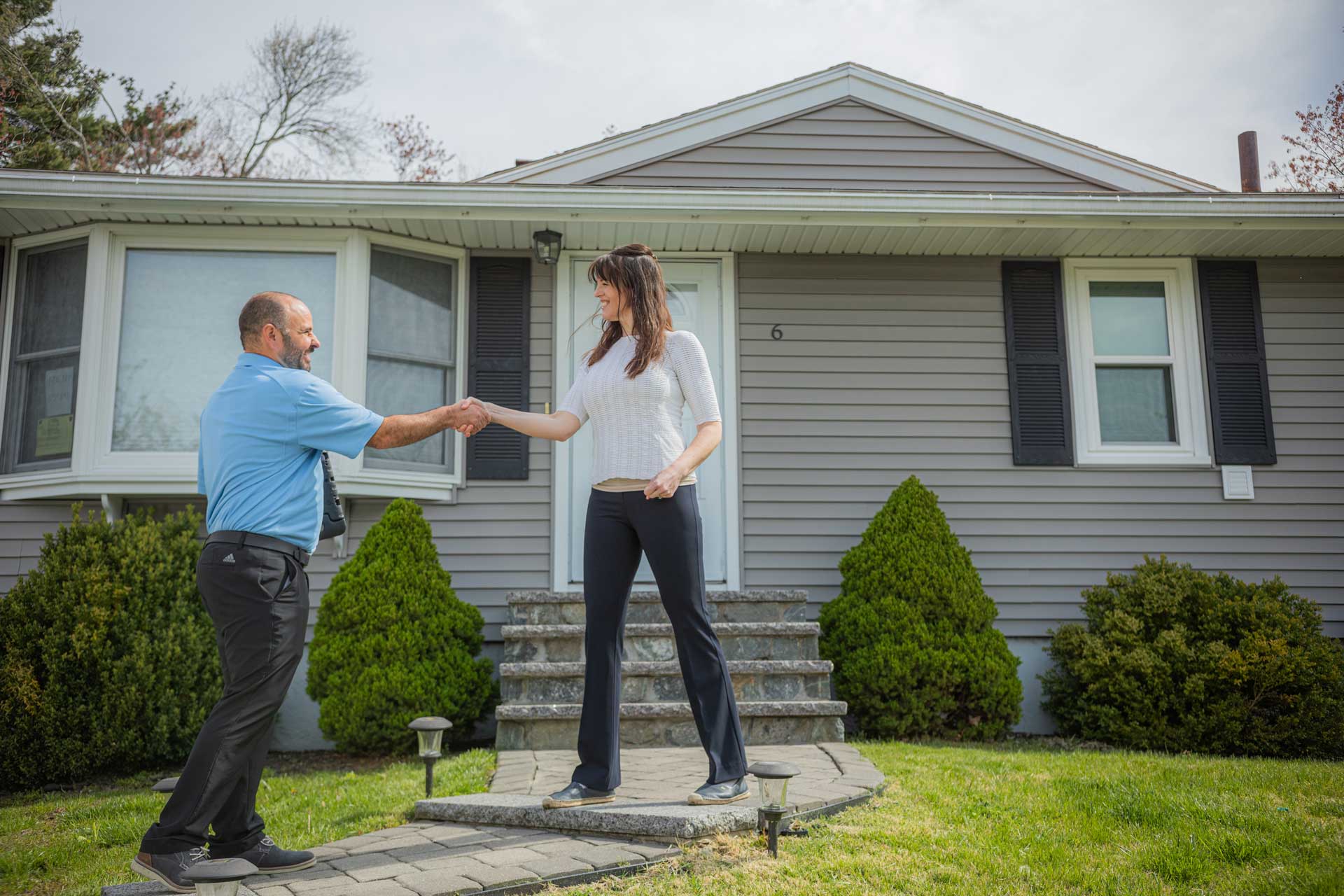A man and woman shake hands outside a gray house with shrubs and steps leading to the entrance.