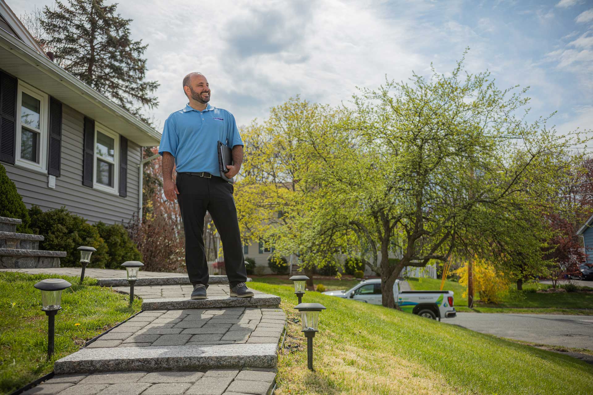 Man in blue shirt stands on walkway outside a house, holding a folder, with trees and a truck in the background.