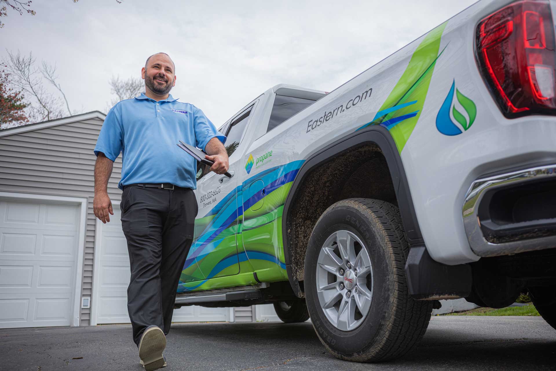 Smiling man in uniform walks beside a branded service truck parked in a residential driveway.