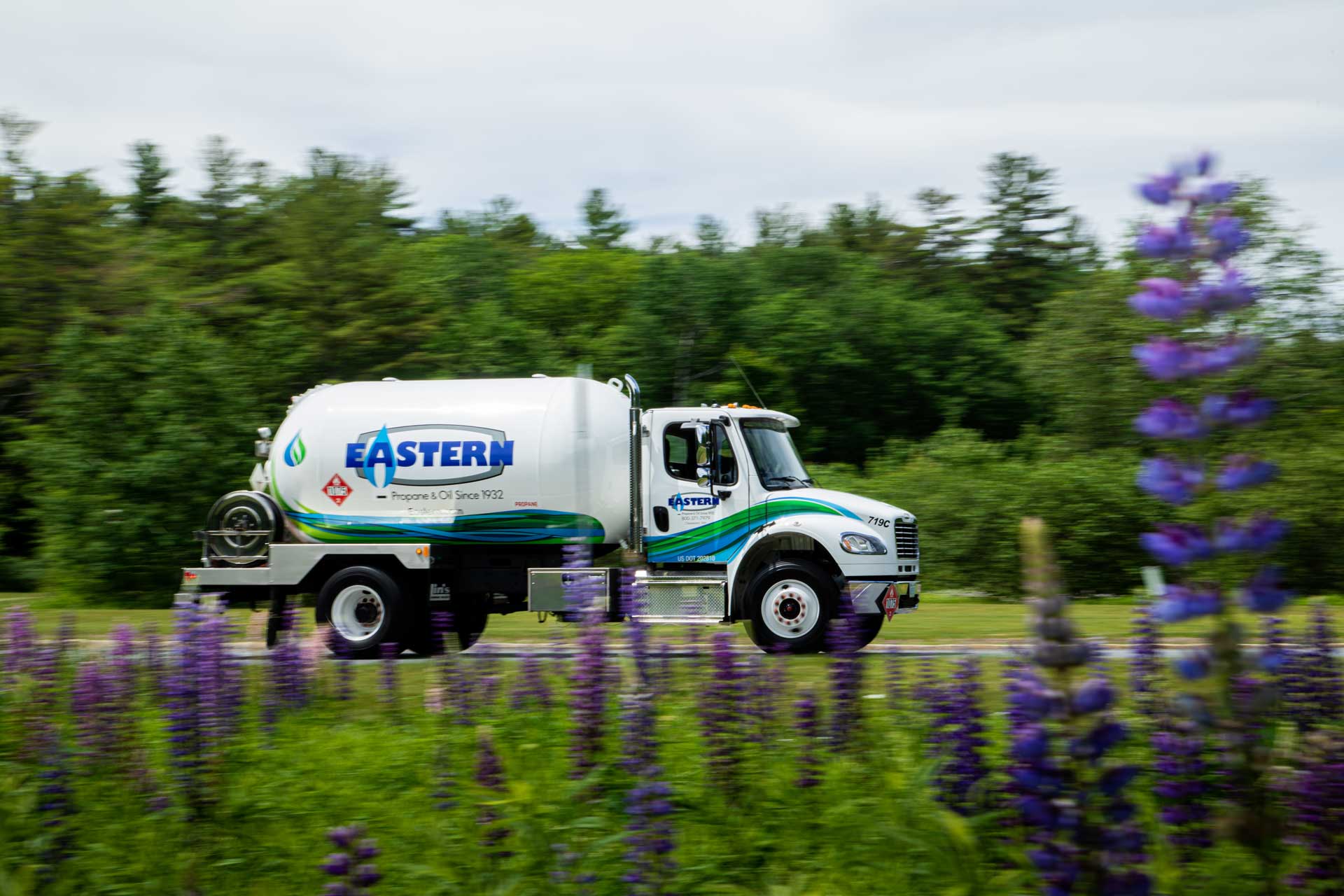 A white Eastern propane truck drives past purple and green wildflowers with trees in the background.