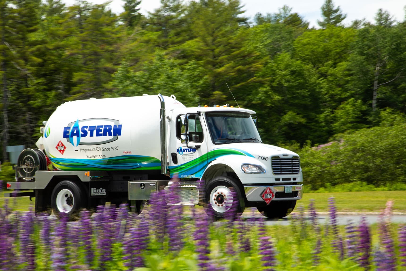A white Eastern propane delivery truck drives past purple flowers and green trees on a sunny day.