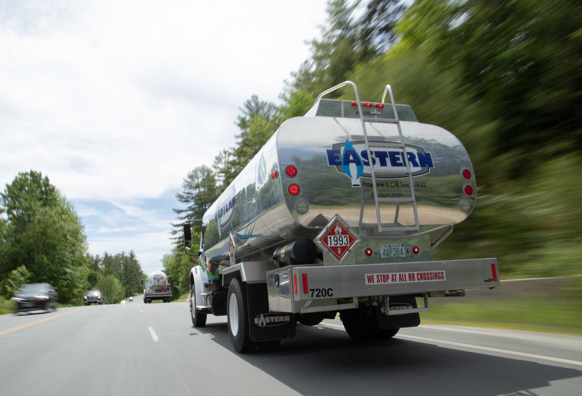 A silver Eastern fuel truck drives down a rural road surrounded by trees on a sunny day.