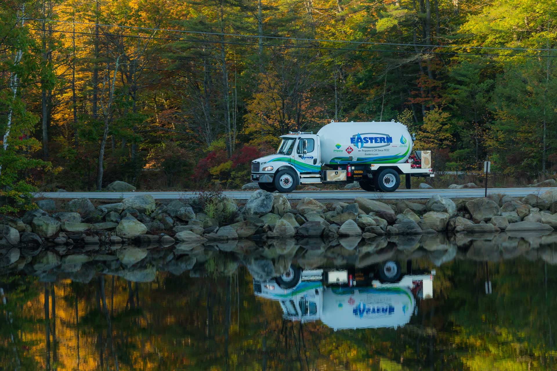 A propane delivery truck drives along a road beside a calm lake with autumn trees reflected in the water.