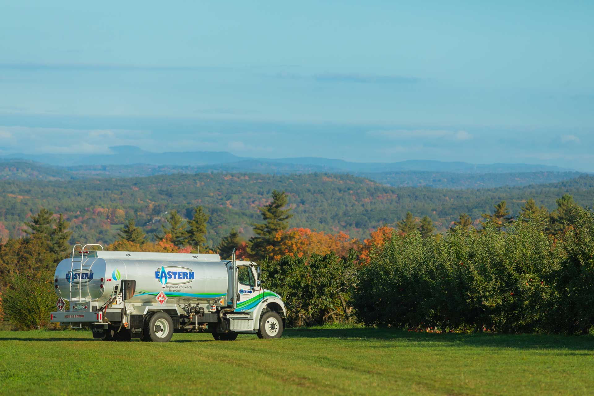 A fuel delivery truck parked on grass with trees and mountains in the background under a blue sky.