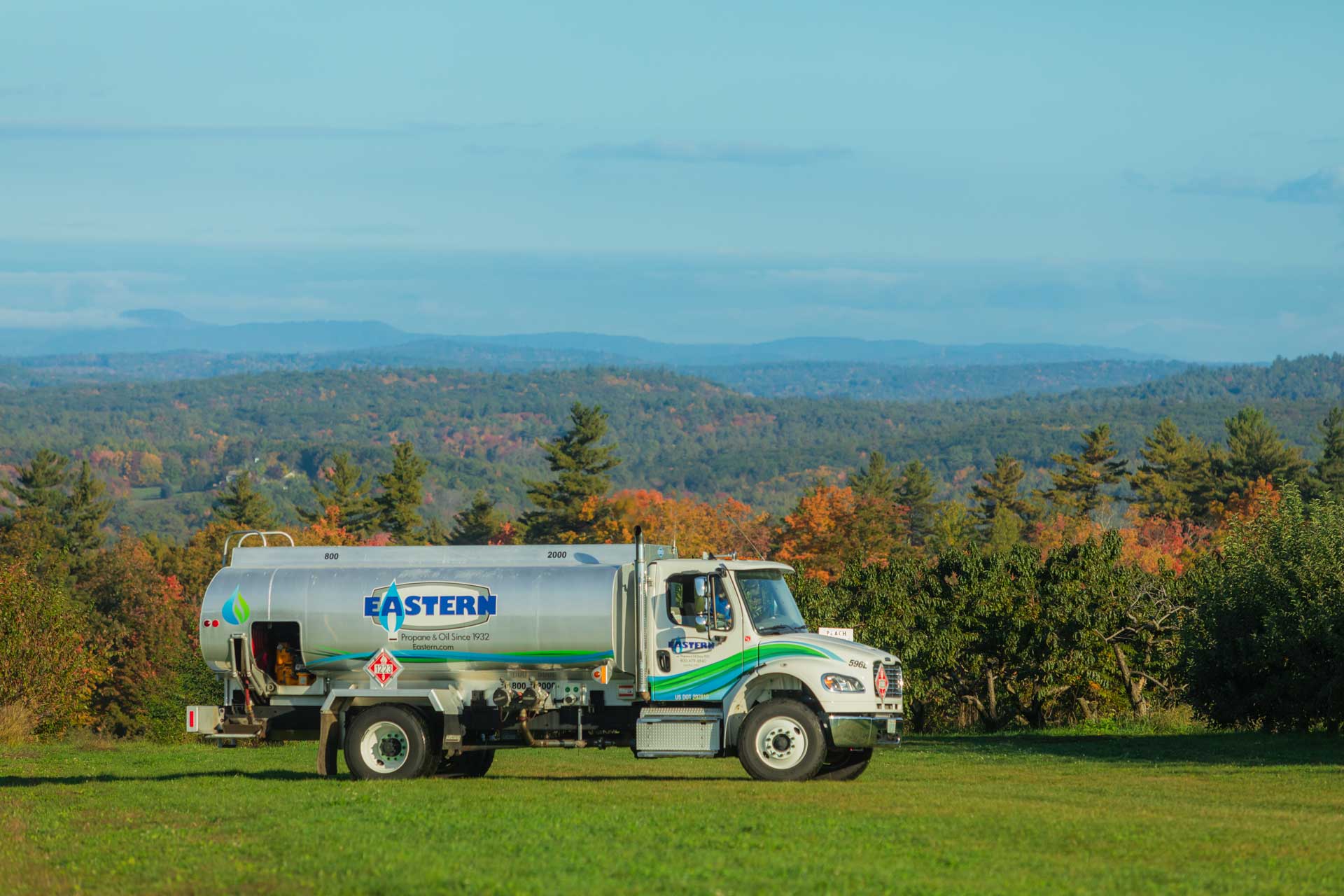 A fuel delivery truck parked on grass with a scenic, forested landscape and hills in the background.