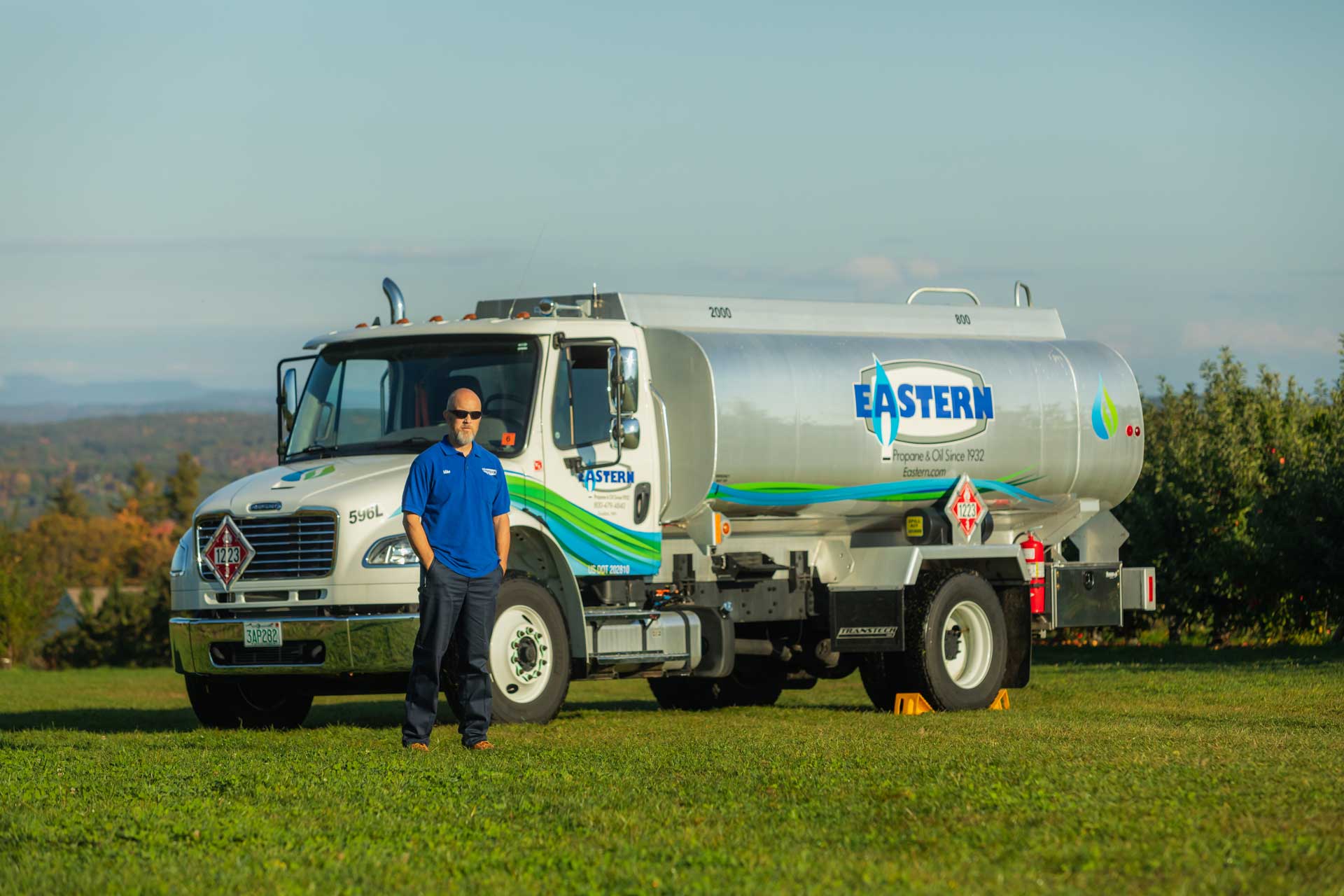 A man in a blue shirt stands in front of an Eastern propane delivery truck on a grassy field.