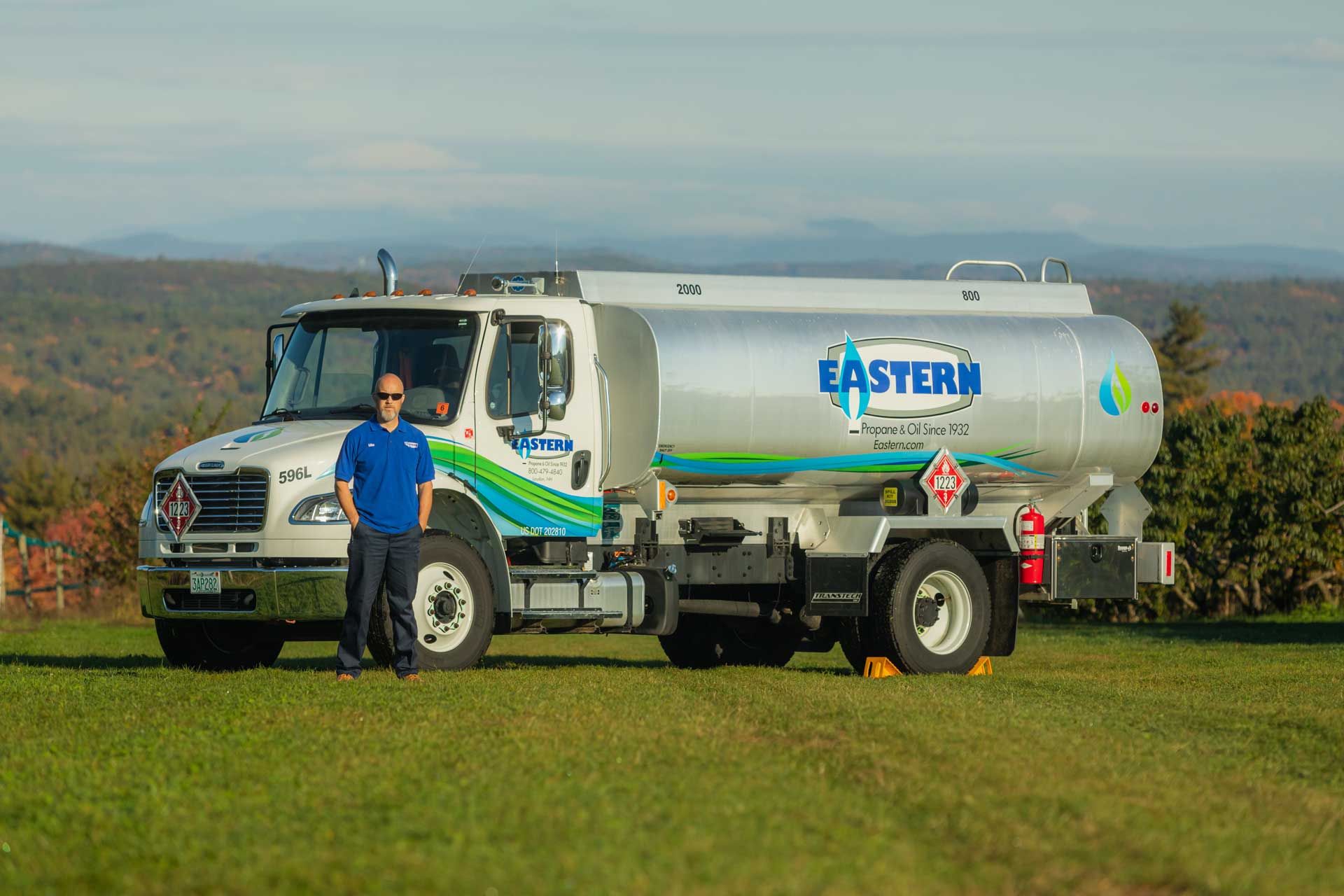 A man in a blue shirt stands beside an Eastern propane delivery truck on a grassy field with hills in the background.