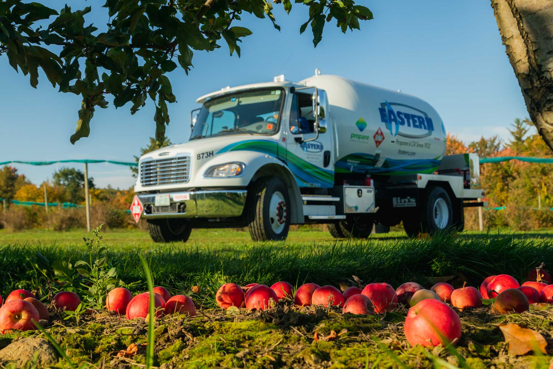 A propane truck is parked in an orchard with red apples scattered on the grass in front.