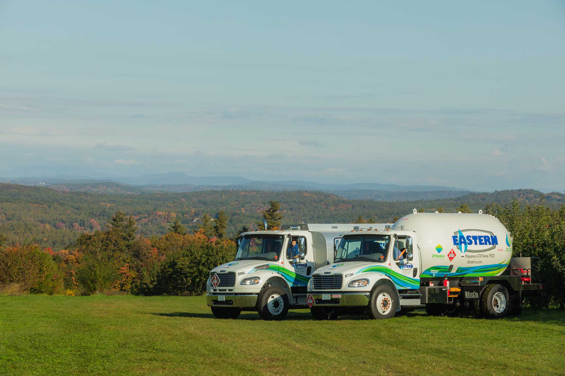 Two Eastern propane trucks parked on grass with a scenic, hilly landscape and trees in the background.
