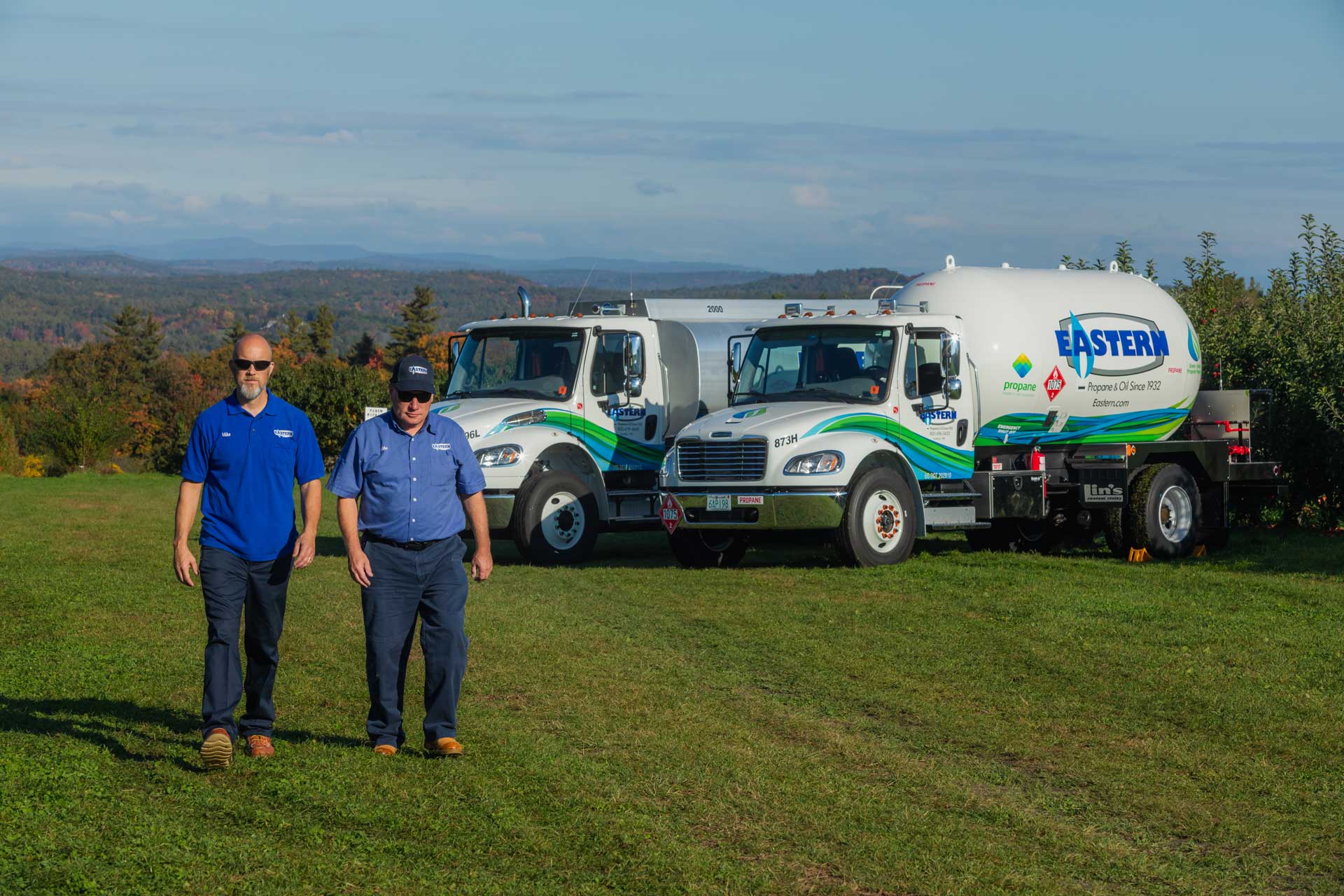 Two men walk on grass near two Eastern propane trucks with a scenic landscape in the background.