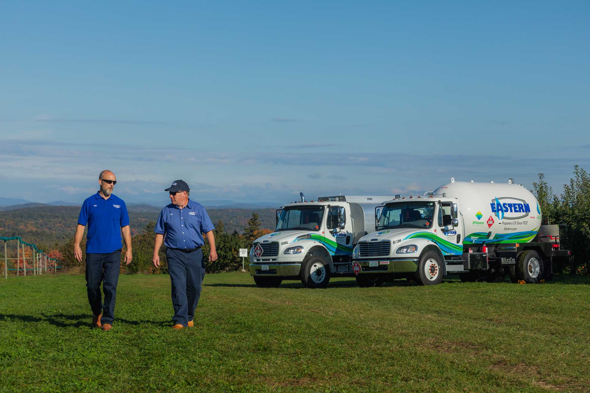 Two men walk on grass near two Eastern propane trucks with hills and trees in the background.