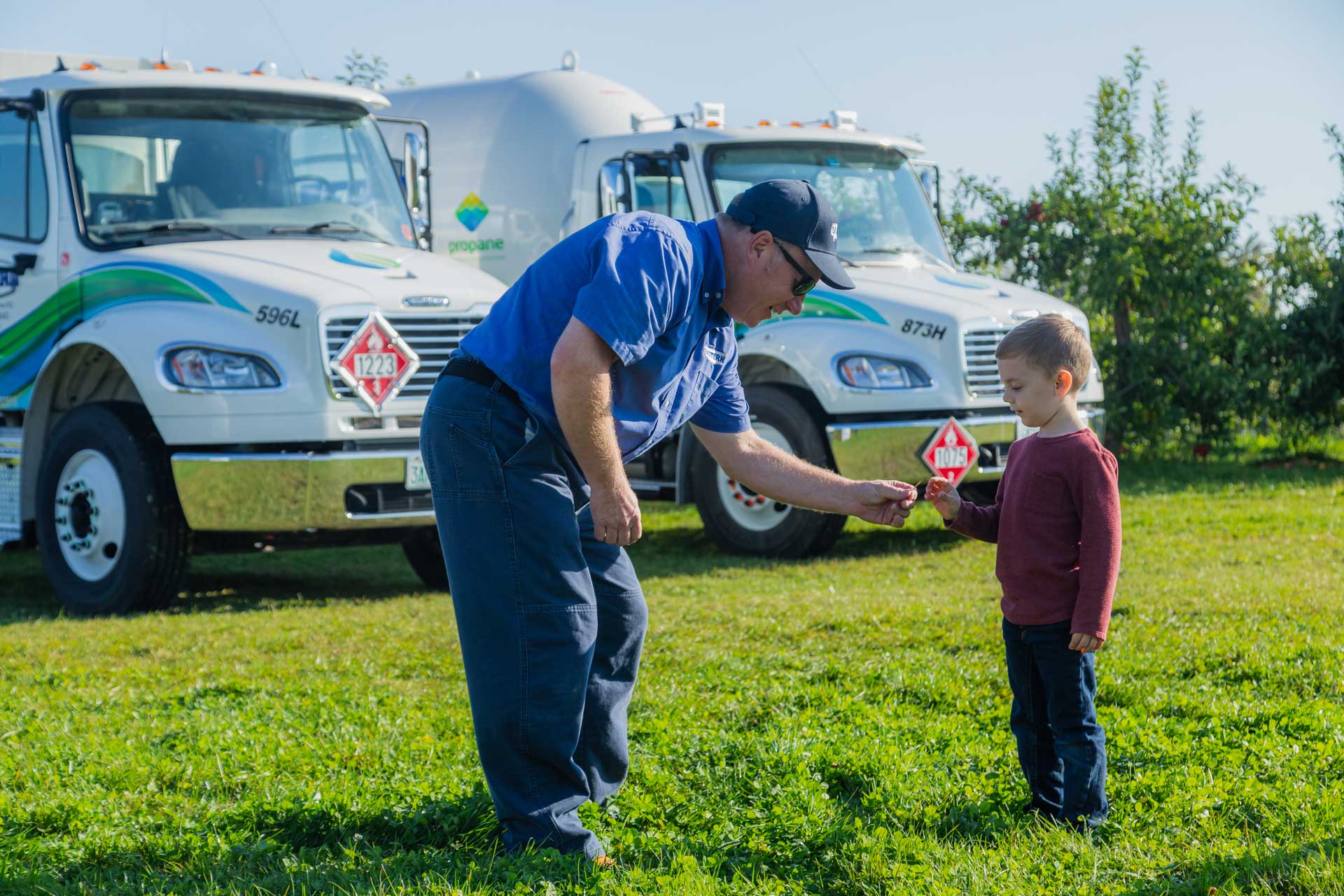 A man hands something to a young boy while standing on grass in front of two propane trucks.