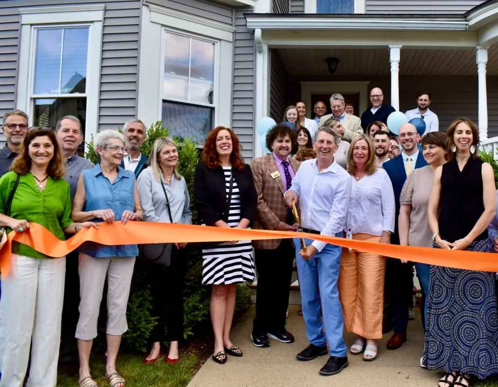 A group of people at a ribbon-cutting ceremony in front of a house, smiling at the camera.
