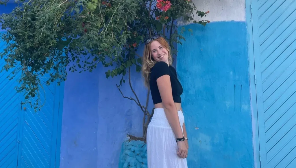 Woman smiling, standing by a blue wall with a leafy tree and red flowers overhead, wearing a black top and white skirt.