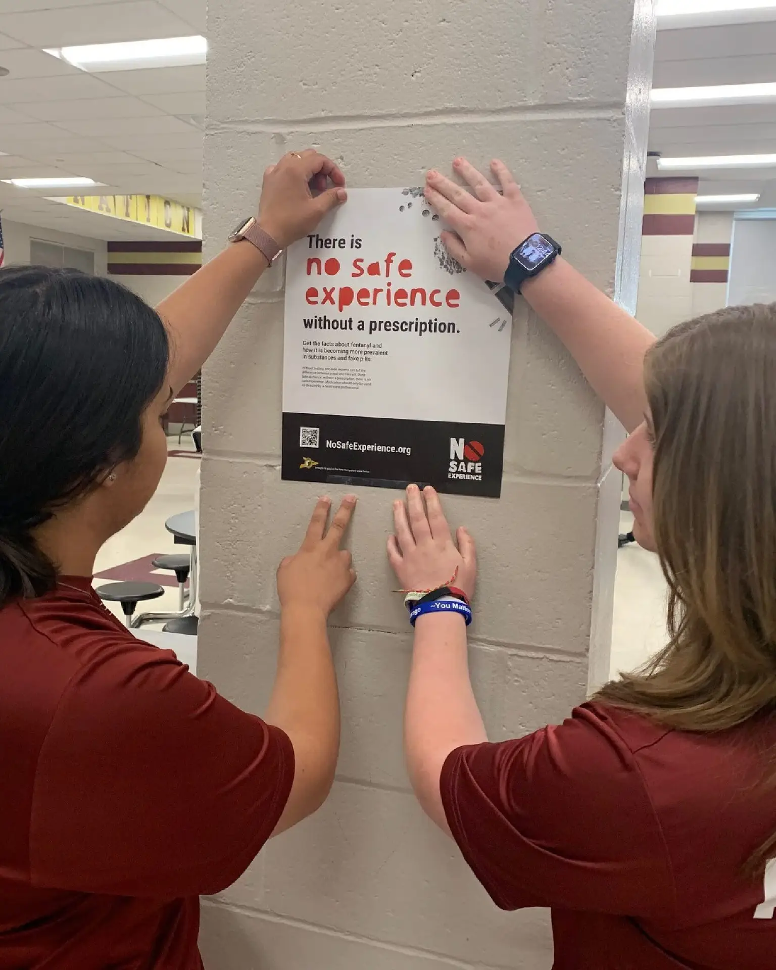Two students hang a No Safe Experience Without a Prescription poster on a school pillar, boosting public awareness about prescription safety.