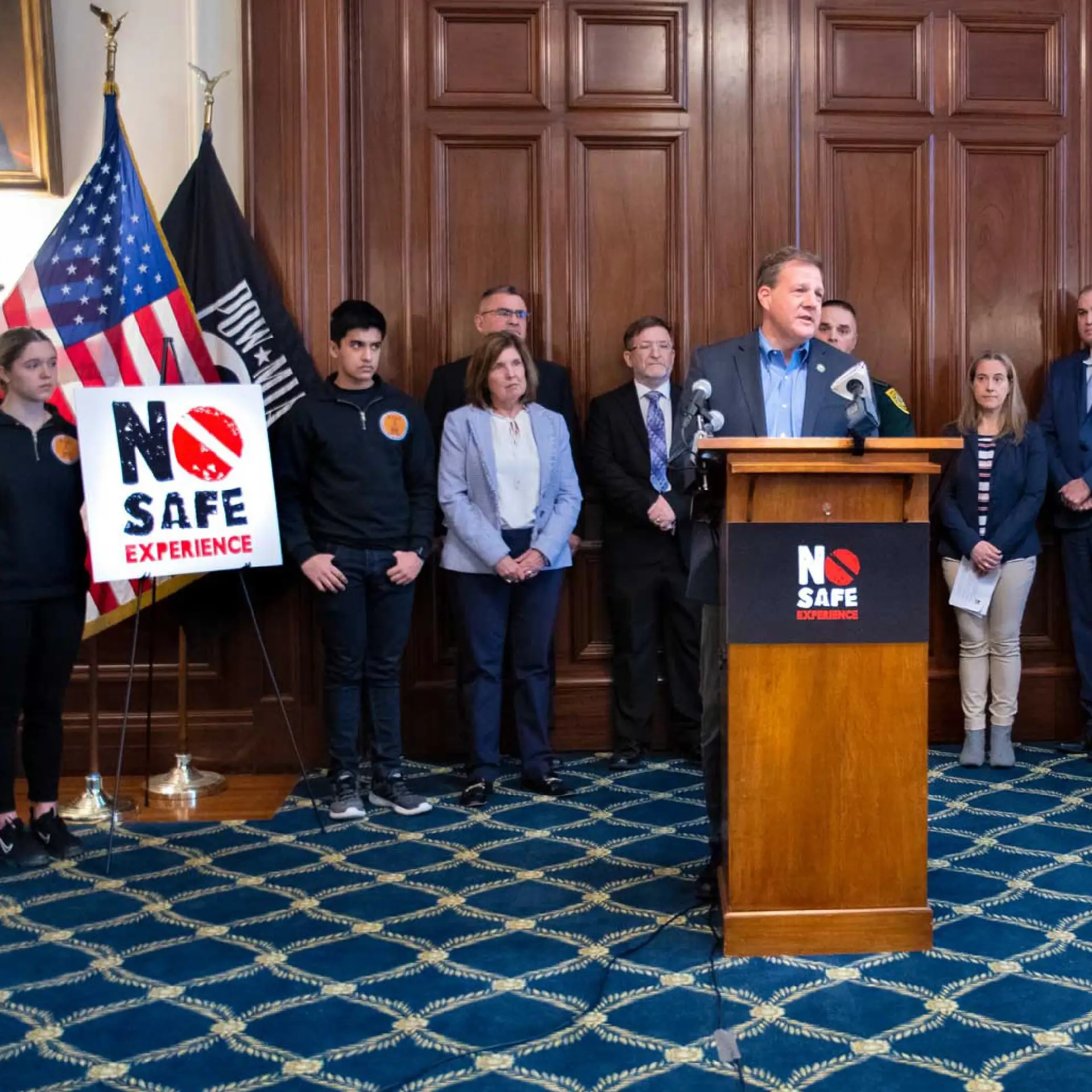 A man speaks at a podium with a No Safe Experience sign, raising public awareness; people stand behind him, U.S. flags at left.
