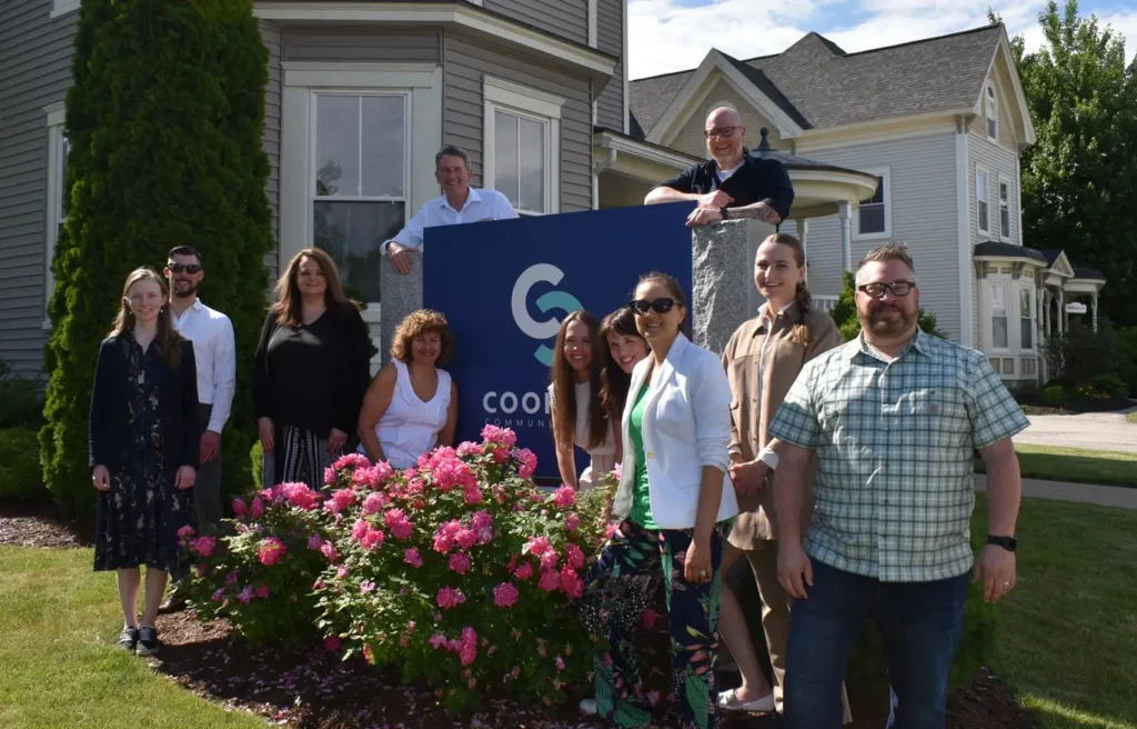 A group of people smiling outside near a blue sign and blooming pink flowers in front of a house.