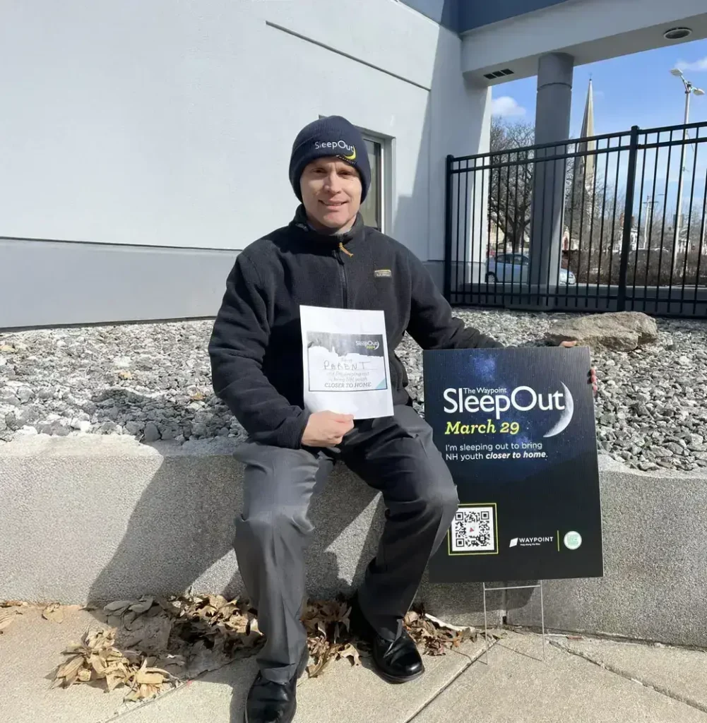 A man in a black jacket and hat sits outside holding a SleepOut sign and a paper in bright sunlight, supporting communications and marketing efforts for the event.