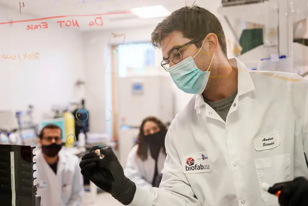A scientist in a lab coat and mask writes on glass while two colleagues watch in a laboratory setting.