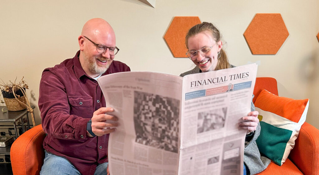 A man and a woman sitting on an orange chair, smiling while reading the Financial Times newspaper together.