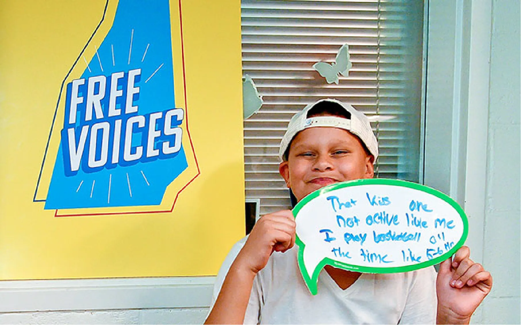 Smiling child in a white cap holds a handwritten sign, standing next to a Free Voices poster.