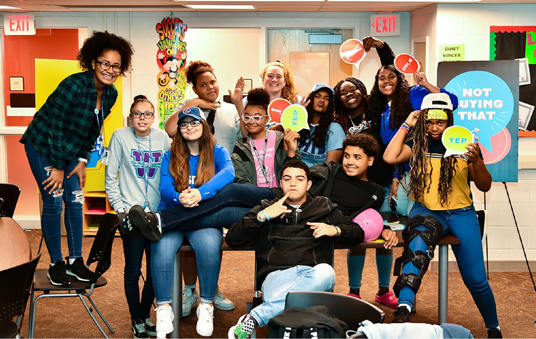 A diverse group of smiling kids and teens pose together, holding colorful signs in a classroom setting.