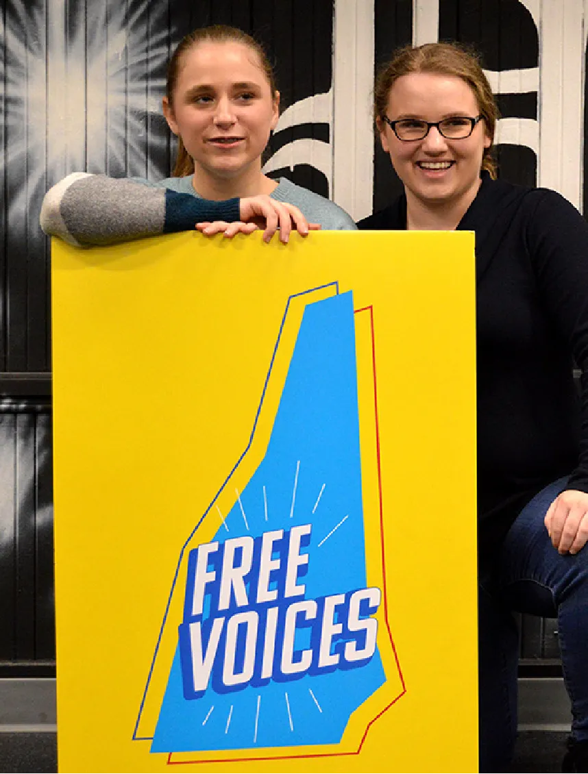 Two women smile behind a bright yellow sign that reads FREE VOICES in bold blue and white letters.