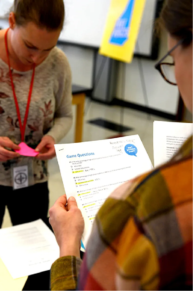 Two people look at printed game questions and notes in a classroom or workshop setting.