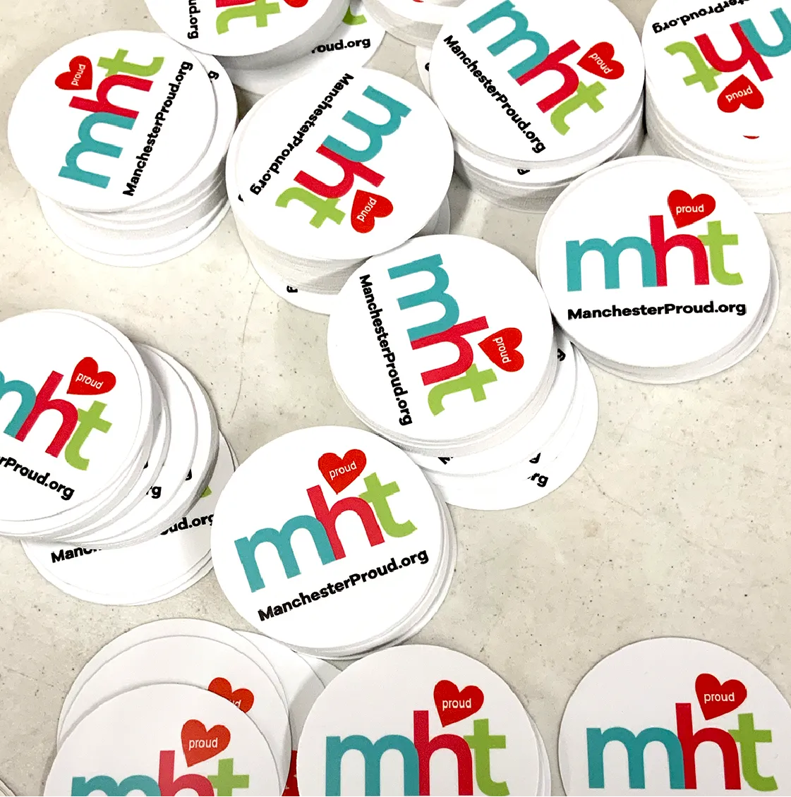 Stacks of round stickers with mht proud and ManchesterProud.org on a white surface.