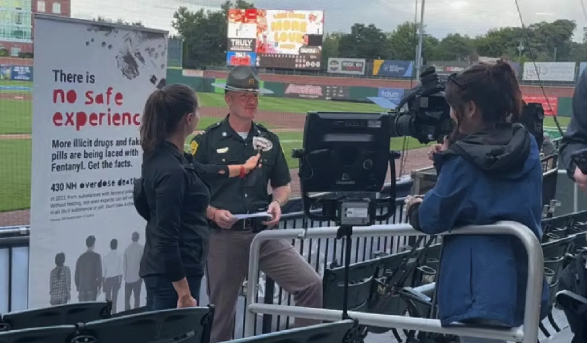 A police officer is interviewed on camera at a stadium near a sign warning about fentanyl dangers.