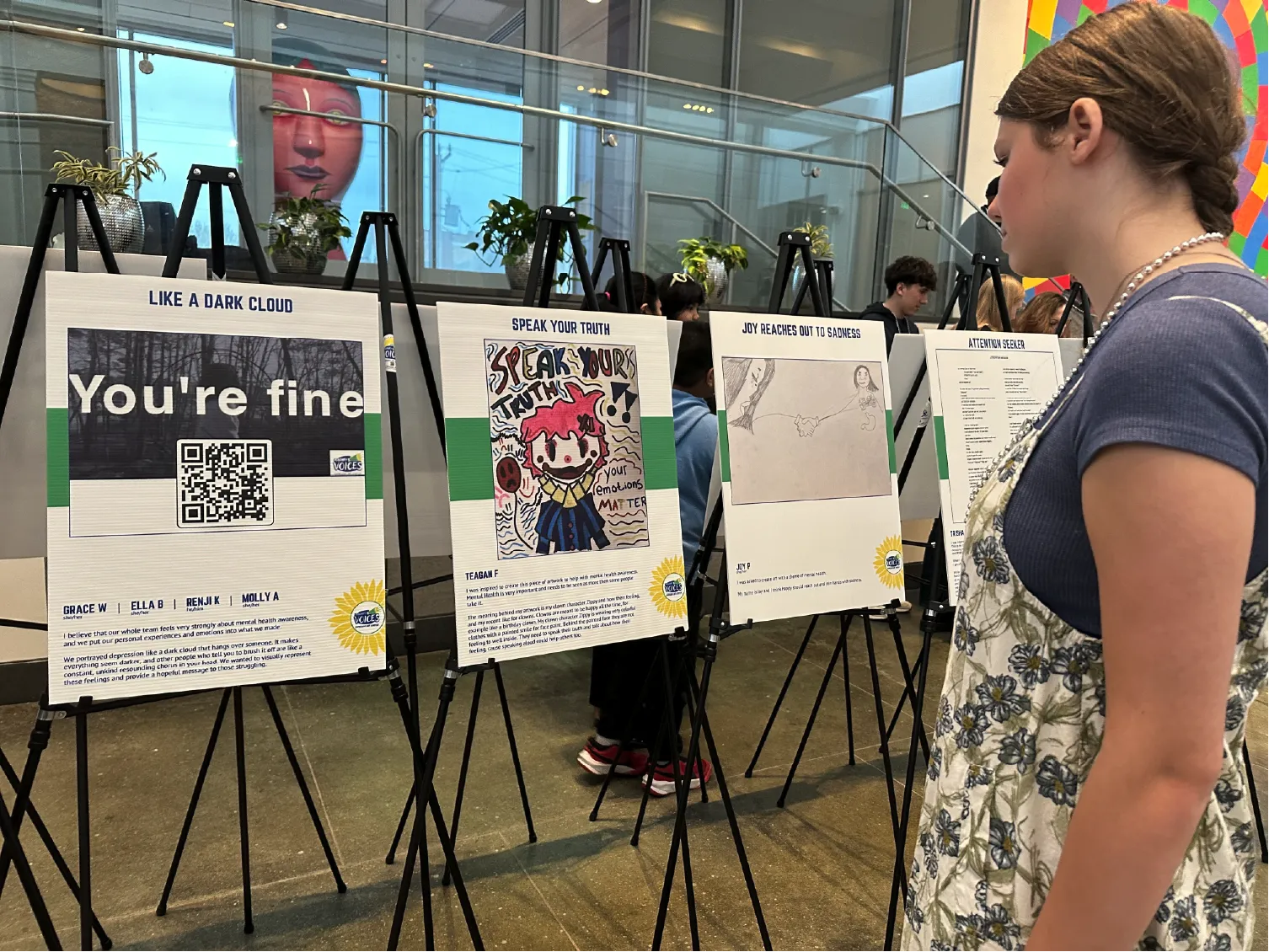 A girl views student artwork and posters displayed on easels in a bright indoor gallery space.
