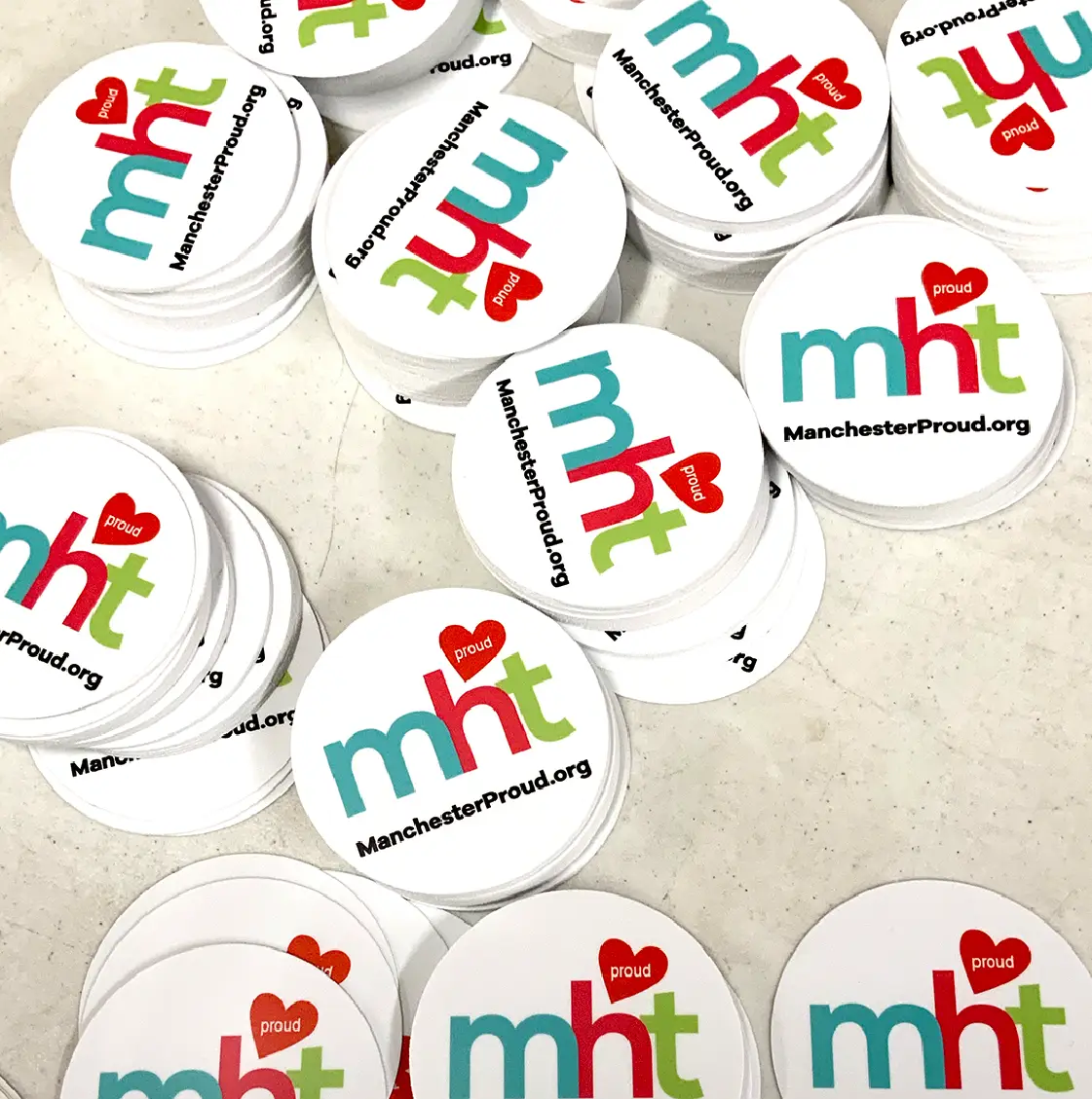 Stacks of white stickers read mht with a red heart labeled proud and ManchesterProud.org below, perfect for Community events or marketing efforts supporting local pride.