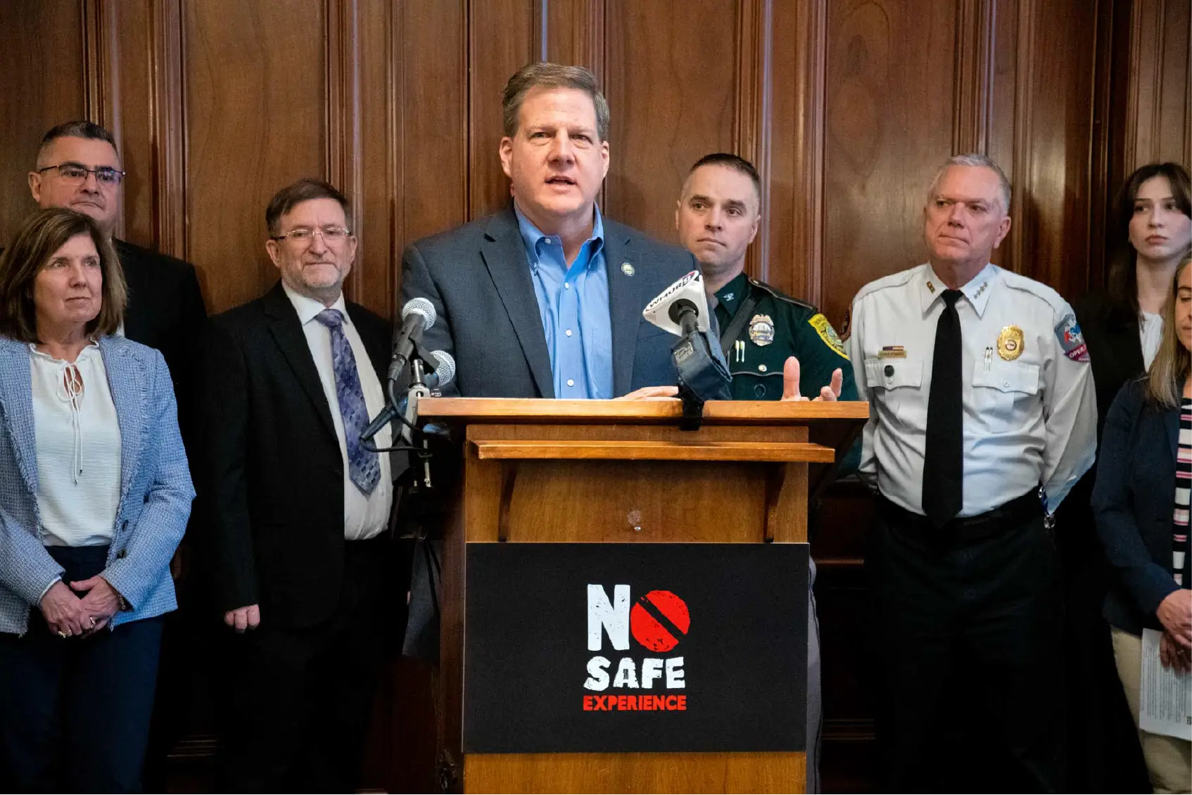 A man speaks at a podium with officials behind him, addressing community concerns; a sign reads No Safe Experience.