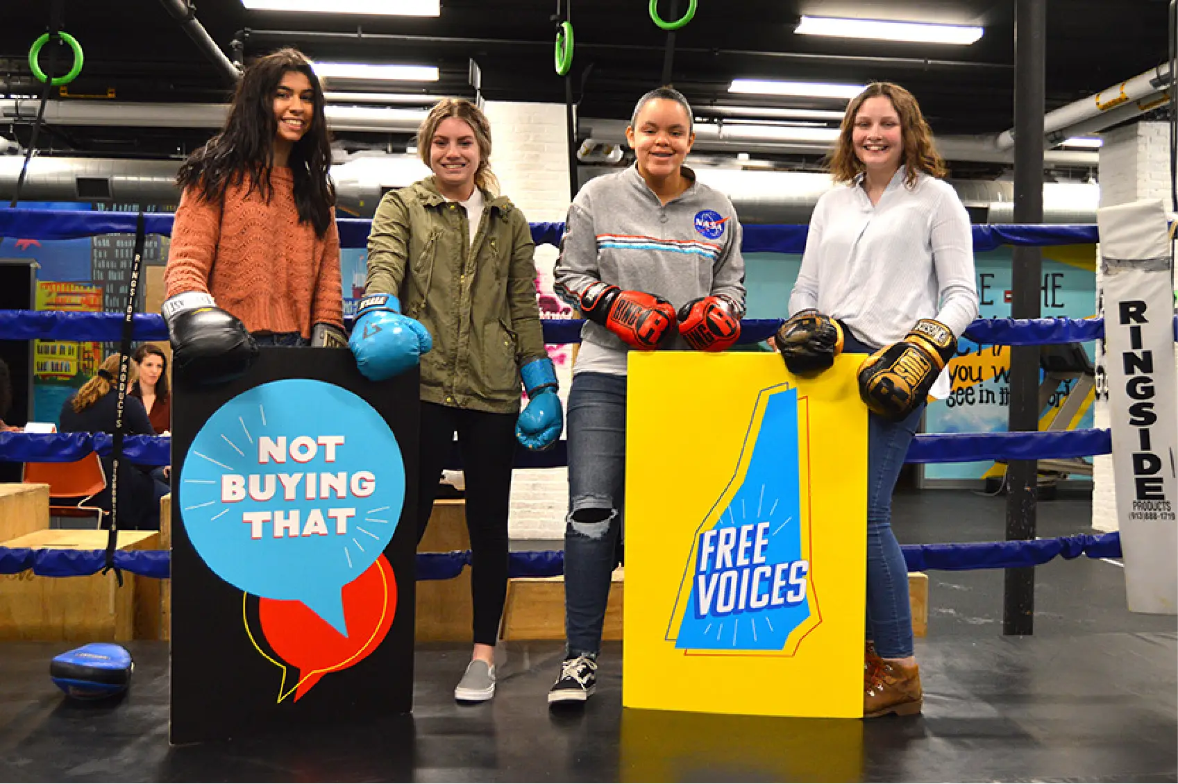 Four young women in a boxing gym, wearing boxing gloves, stand united for Community empowerment as they hold signs reading “Not Buying That” and “Free Voices”—a bold statement against manipulative marketing.