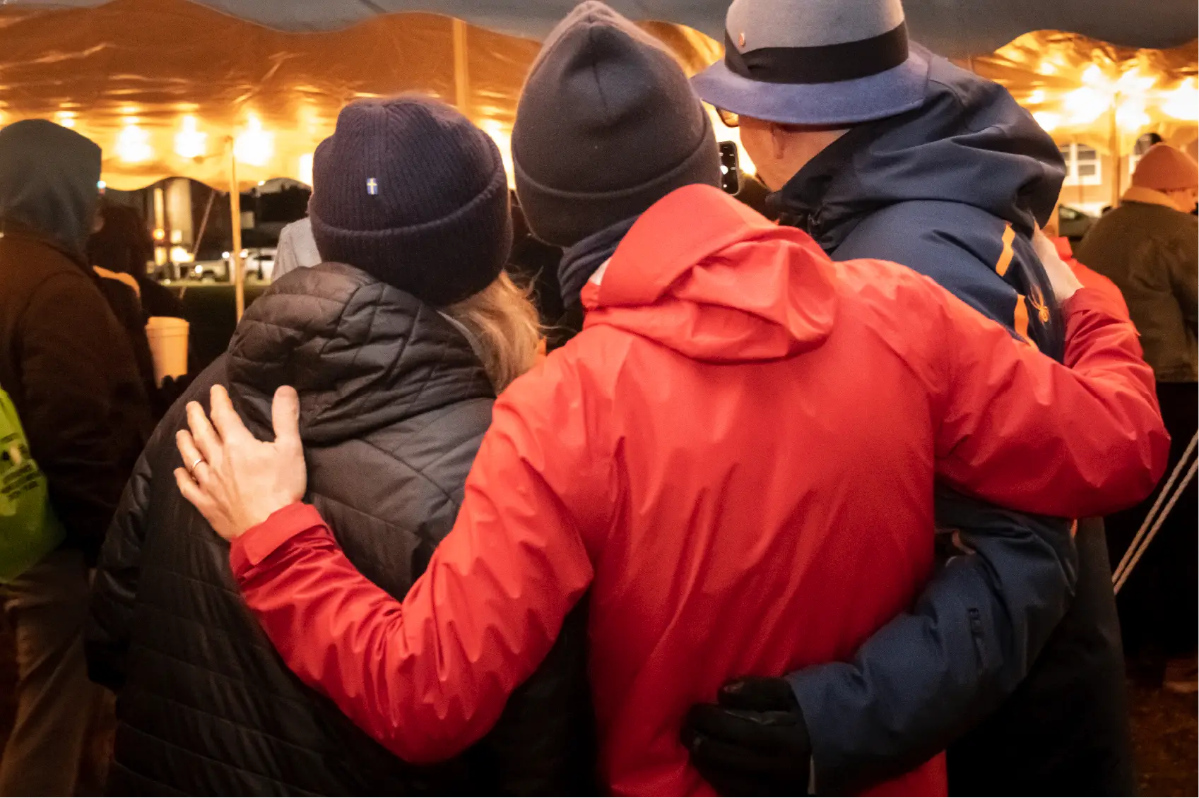 Three people in winter jackets and hats stand close together under a tent, arms around each other, showcasing the warmth of community.